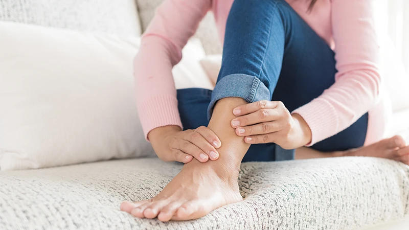 A woman in jeans and a pink shirt massages her ankle injury while sitting on a couch.