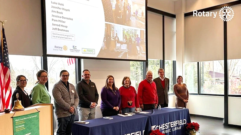 group of Rotary Club members standing behind a table during a meeting at Chesterfield Public Library, with a presentation screen in the background.