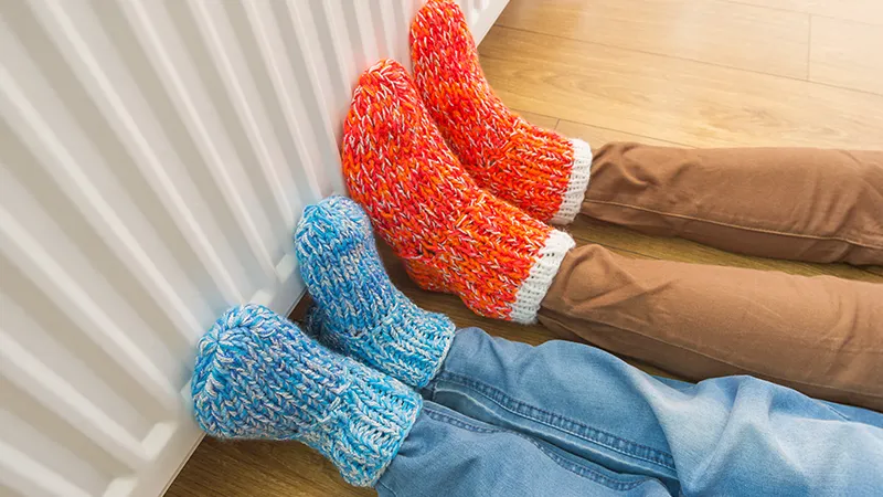 Two people relaxing with their feet up against a white radiator, wearing thick knitted socks, one pair bright red and white, the other blue and white.